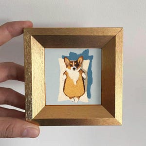 May include: A framed artwork featuring a corgi dog resting on a pillow. The corgi has brown, white, and tan fur. The artwork is set against a light blue background and framed in a gold-colored wooden frame.