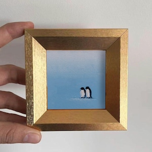 May include: A small framed painting of two penguins on a light blue background. The penguins are black and white, and the frame is gold-coloured. The painting is held by a hand.