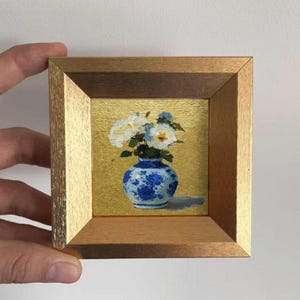 Puede incluir: Una pintura en miniatura enmarcada de flores blancas en un jarrón azul y blanco. La obra de arte está sobre un fondo dorado y enmarcada en un marco de madera. La pintura está sostenida por una mano.