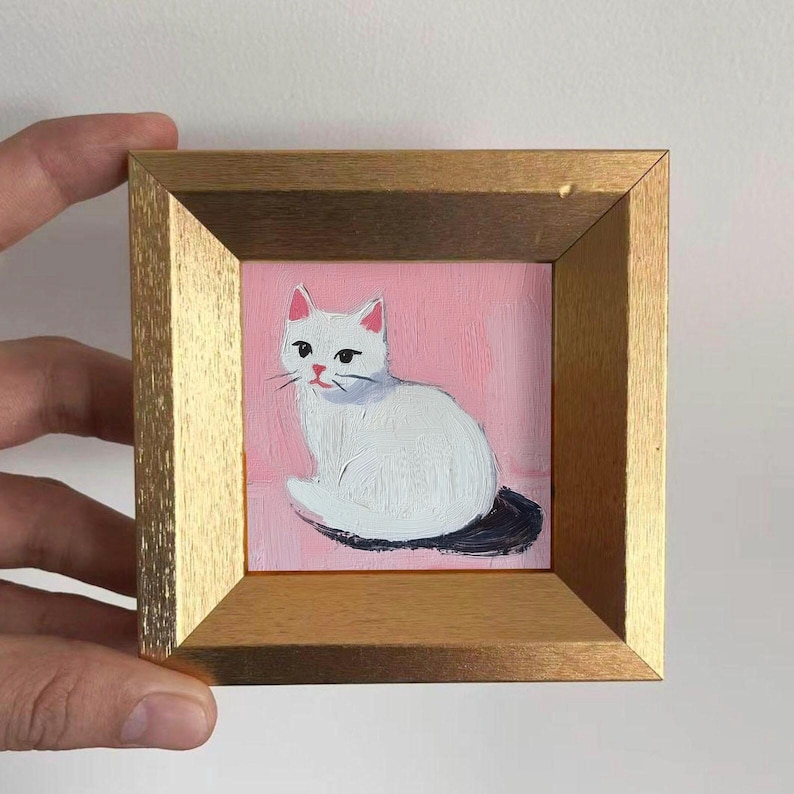 May include: A small, square painting of a white cat with pink ears and a black tail, set against a pink background. The artwork is framed in a gold-colored wooden frame, held by a hand.