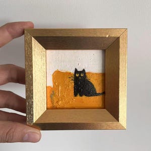 May include: A small, framed painting featuring a black cat with yellow eyes. The cat is set against a textured orange and white background. The frame is a light gold color, adding a touch of elegance to the artwork.