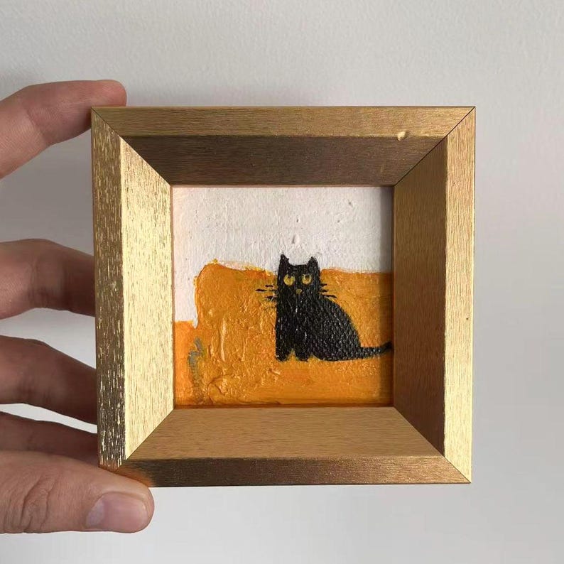May include: A small, framed painting featuring a black cat with yellow eyes. The cat is set against a textured orange and white background. The frame is a light gold color, adding a touch of elegance to the artwork.