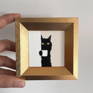 Puede incluir: Pintura enmarcada que representa un gato negro con ojos amarillos sosteniendo una taza blanca. El fondo se divide entre blanco texturizado y beige. El marco dorado realza la estética de la obra de arte. El gato parece estar escondiéndose detrás de una pared.