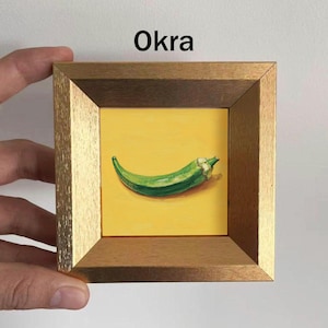 May include: A framed painting of a green okra pod on a yellow background. The artwork is set within a gold-toned wooden frame, with the word "Okra" displayed at the top.