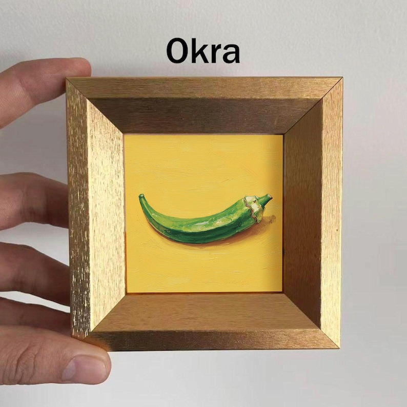May include: A framed painting of a green okra pod on a yellow background. The artwork is set within a gold-toned wooden frame, with the word "Okra" displayed at the top.