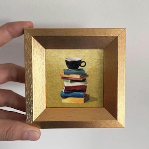Puede incluir: Una pintura en miniatura enmarcada representa una pila de libros coloridos coronada por una taza de café negra. La obra de arte está sobre un fondo dorado y enmarcada en un marco de madera, sostenida por una mano.