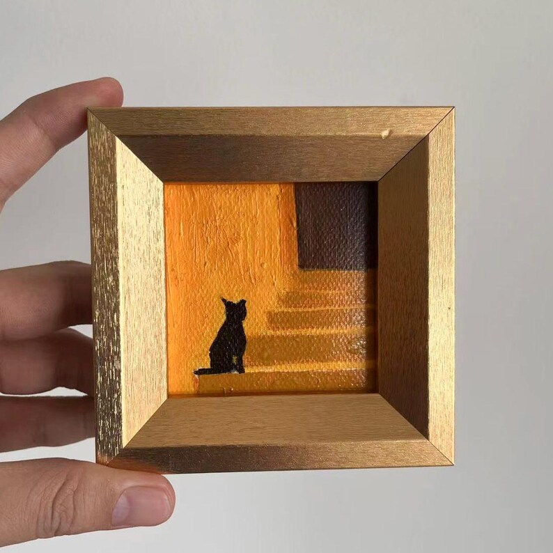 May include: Small framed painting featuring a black cat silhouette on a staircase. The artwork has a warm color palette with orange and brown tones. The frame is a gold color.