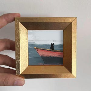 Op de afbeelding: Een ingelijst miniatuurschilderij toont een rode boot op het water met een zwart katten silhouet. Het kunstwerk staat tegen een achtergrond van een grijze lucht en water, omlijst door een goudkleurige houten lijst.