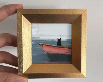 May include: A framed miniature painting depicts a red boat on water with a black cat silhouette. The artwork is set against a backdrop of a gray sky and water, framed in a gold-colored wooden frame.