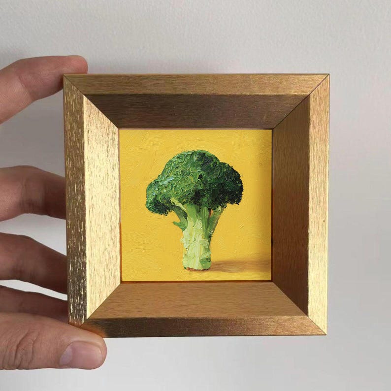 May include: A small, framed painting of a broccoli floret. The broccoli is rendered in shades of green and sits against a bright yellow background. The frame is a warm gold color, adding a touch of elegance to the artwork.