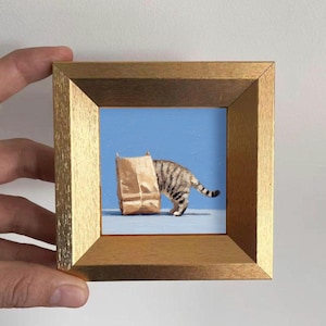May include: A framed painting depicts a tabby cat with its head inside a brown paper bag. The cat is standing on a light blue surface against a solid blue background. The frame is a light gold color.