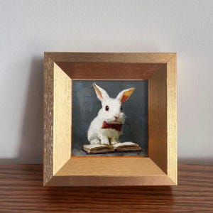 Peut inclure: Peinture encadrée d'un lapin blanc portant un nœud papillon rouge, assis sur un livre ouvert. L'œuvre est encadrée d'or et présente un fond gris foncé. Le lapin a les oreilles roses et est le sujet principal de la peinture.