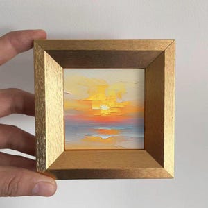 Könnte beinhalten: Gerahmtes Miniatur-Ölgemälde, das einen Sonnenuntergang darstellt. Das Kunstwerk verwendet eine lebendige Farbpalette aus Gelb, Orange und Blau. Das Gemälde ist in einem goldfarbenen Holzrahmen gefasst und verleiht der Miniaturlandschaft Eleganz.