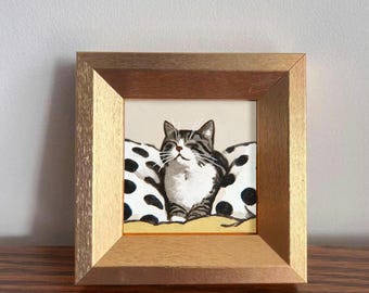 May include: A framed painting of a cat resting on pillows. The cat has gray and white stripes and is smiling. The pillows are white with black polka dots. The frame is a light brown color.
