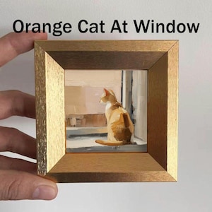 May include: A framed painting titled "Orange Cat At Window" depicts an orange and white cat gazing out a window. The artwork is set within a gold-colored wooden frame. The painting's style is impressionistic, with visible brushstrokes.