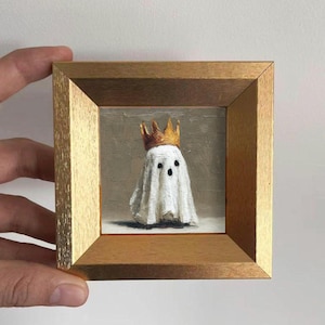 May include: A small square painting in a gold-colored frame featuring a white ghost figure wearing a gold crown. The ghost has two black eyes and a small black mouth, set against a neutral gray background with visible brushstroke textures.