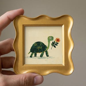 Op de afbeelding: Een klein vierkant schilderij in een gouden golvende lijst met een groene schildpad met een donkergroen schild die een oranje bloem in de bek houdt. Het schilderij heeft een crèmekleurige achtergrond en zichtbare penseelstreken in het werk.