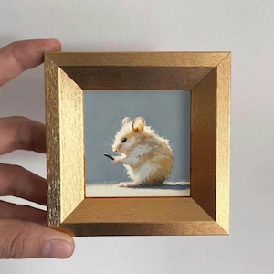 Könnte beinhalten: Gerahmtes Miniaturgemälde eines Hamsters, der ein kleines schwarzes Objekt hält. Der Hamster ist in Beige- und Weißtönen vor einem hellblauen Hintergrund dargestellt. Der Rahmen ist goldfarben und verleiht dem Kunstwerk Wärme.