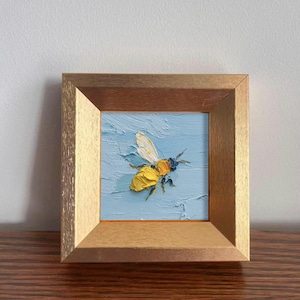 May include: A small, framed painting of a bee. The bee is rendered in thick, textured yellow and black paint, set against a light blue background. The frame is a warm, light brown color, adding a natural touch to the artwork.