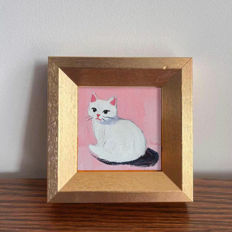 May include: A framed painting of a white cat with pink ears and a black tail, set against a pink background. The artwork is encased in a gold-colored wooden frame, creating a decorative piece.