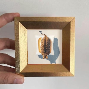Könnte beinhalten: Ein gerahmtes Kunstwerk mit einer Katze mit braunen und schwarzen Streifen, die auf einem weißen Kissen und einer Toastscheibe liegt. Das Kunstwerk befindet sich in einem goldfarbenen Holzrahmen, der von einer Hand gehalten wird.