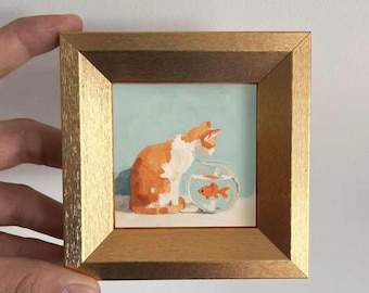 May include: A framed painting depicts a ginger and white cat gazing at a goldfish in a glass bowl. The artwork is set against a light blue background and framed in a gold-colored wooden frame.