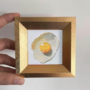 Könnte beinhalten: Ein kleines, gerahmtes Gemälde eines Spiegeleis. Das Kunstwerk zeigt ein leuchtend gelbes Eigelb und ein weißes und graues Eiweiß mit sichtbaren Pinselstrichen. Der Rahmen ist hellbraun.