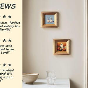 May include: Two small framed paintings are displayed on a wall. One shows two white birds on a branch against a blue sky. The other depicts a ghost, pumpkins, and a candle. The frames are gold-coloured.