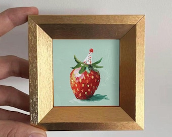 May include: A small, square painting in a gold frame. The artwork depicts a red strawberry wearing a party hat against a light teal background. The strawberry has green leaves and a shadow.