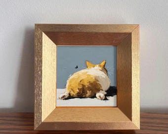 May include: A framed painting of a cat, viewed from the rear, with orange and white fur, resting on a white surface against a blue background. A small insect flies nearby. The painting is in a gold-colored wooden frame.