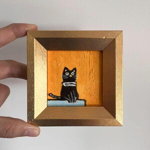May include: A small, square painting of a black cat with a white scarf, set against an orange background. The artwork is framed in a gold-colored wooden frame. The cat is sitting on a light blue surface.