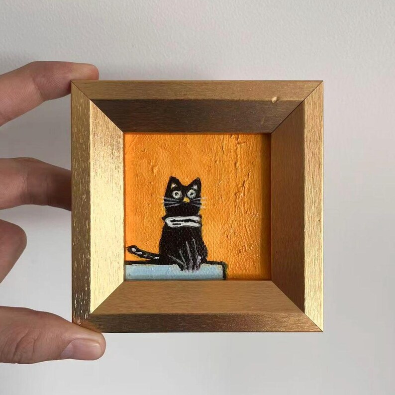 May include: A small, square painting of a black cat with a white scarf, set against an orange background. The artwork is framed in a gold-colored wooden frame. The cat is sitting on a light blue surface.