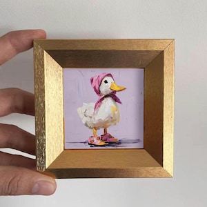 Puede incluir: Pintura enmarcada de un pato blanco con un gorro, bufanda y zapatos rosas. El pato tiene un pico amarillo y está sobre un fondo lavanda. El marco es de color dorado.