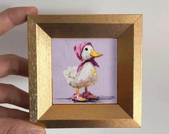May include: A framed painting of a white duck wearing a pink bonnet, scarf, and shoes. The duck has a yellow beak and is set against a lavender background. The frame is gold-colored.