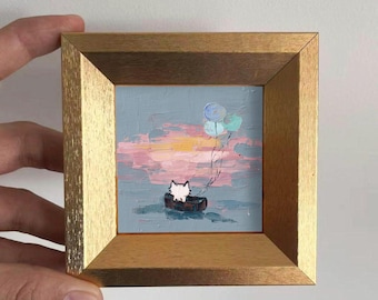 May include: A small, square painting in a gold frame. The artwork depicts a white cat in a boat, with balloons floating above. The background features a pastel sky with pink, yellow, and blue hues, creating a whimsical scene.