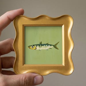 Può includere: Un piccolo dipinto quadrato di un singolo pesce centrato su uno sfondo verde chiaro, racchiuso in una cornice decorativa color oro con un bordo ondulato e smerlato. Il pesce è raffigurato in tonalità di blu, verde e giallo con un occhio.