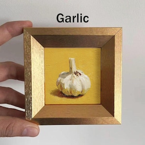 May include: A framed painting of a garlic bulb on a yellow background. The garlic is white with a textured surface and a green stem. The frame is gold and brown, and the word "Garlic" is written in black.