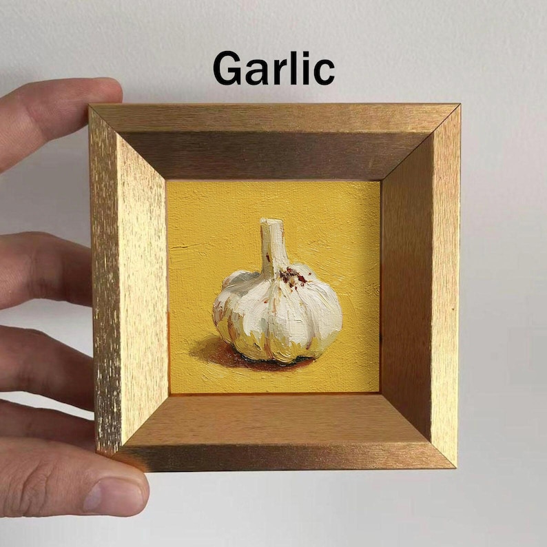 May include: A framed painting of a garlic bulb on a yellow background. The garlic is white with a textured surface and a green stem. The frame is gold and brown, and the word "Garlic" is written in black.