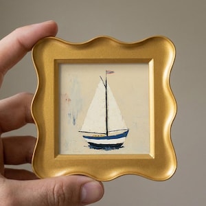 May include: A small square painting of a sailboat with a white sail and a blue and white hull, featuring a tiny flag at the top of the mast. The artwork is encased in a decorative gold frame with a wavy, scalloped edge and a smooth matte finish.