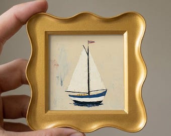 May include: A small square painting of a sailboat with a white sail and a blue and white hull, featuring a tiny flag at the top of the mast. The artwork is encased in a decorative, wavy-edged gold frame with a smooth, metallic finish.