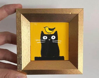 Puede incluir: Pintura enmarcada que representa un gato negro con un pájaro amarillo en la cabeza, sobre un fondo amarillo. La obra de arte está en un marco de madera con un borde interior dorado. El gato tiene bigotes blancos y ojos grandes y blancos.