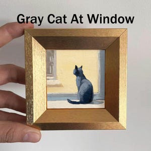 May include: A framed painting titled "Gray Cat At Window" depicts a gray cat gazing out a window. The artwork is set in a gold-toned wooden frame. The cat is painted in shades of gray, and the window has a yellow background.