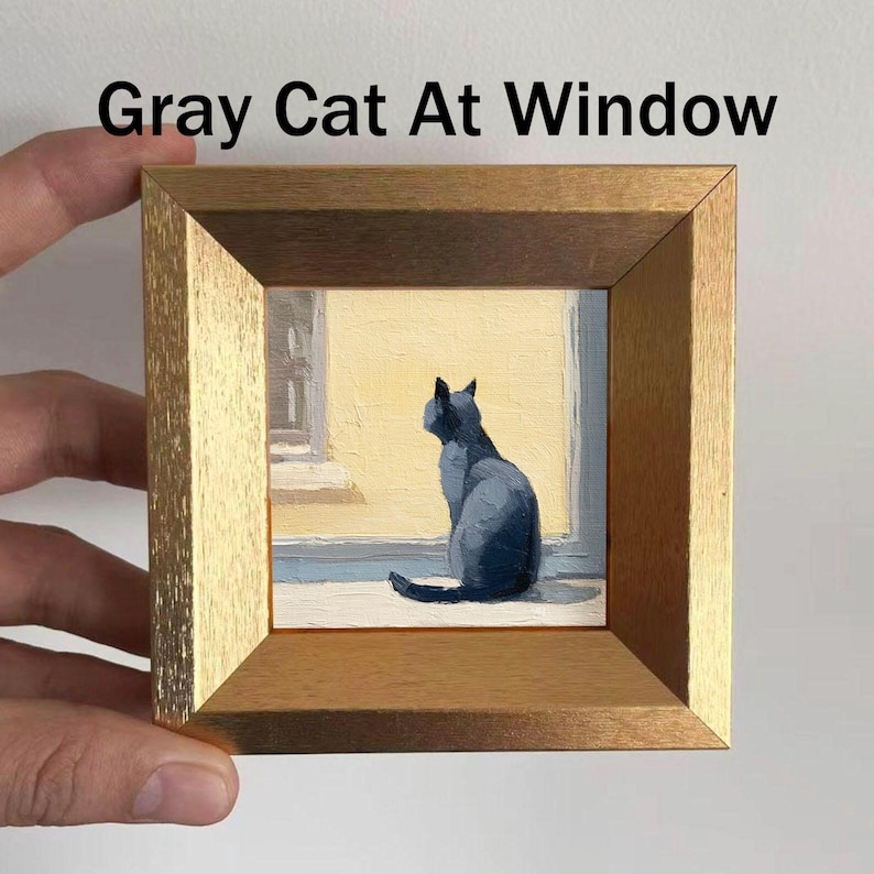 May include: A framed painting titled "Gray Cat At Window" depicts a gray cat gazing out a window. The artwork is set in a gold-toned wooden frame. The cat is painted in shades of gray, and the window has a yellow background.
