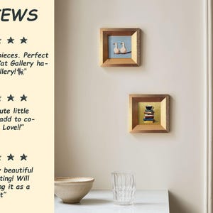 May include: Two small framed paintings with gold-colored frames hang on a cream-colored wall. One painting depicts two white birds on a branch, the other shows a stack of books and a teacup. A review section is visible.