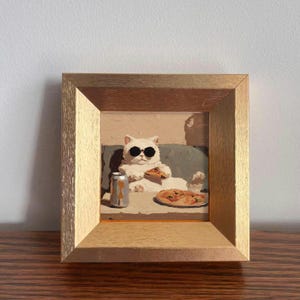 Può includere: Un'opera d'arte incorniciata raffigura un gatto bianco con occhiali da sole, seduto su un divano, che si gode una pizza e una bibita. Il gatto tiene in mano una fetta di pizza. Una pizza intera e una lattina di bibita sono sul tavolo. La cornice è dorata.