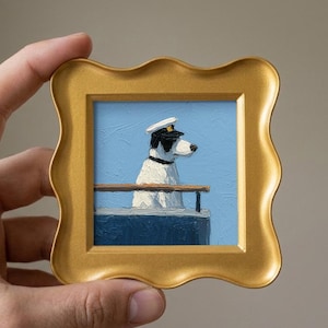 May include: A small, framed painting of a dog wearing a sailor's hat, standing on a ship's deck. The painting has a gold frame with a wavy edge. The dog is white with black markings, set against a blue sky.