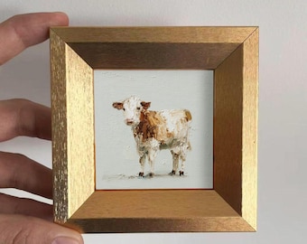 May include: A small framed painting of a cow. The cow is depicted with white and brown textured brushstrokes, standing on a light blue background. The frame is a gold color. The painting is held by a person's hand.