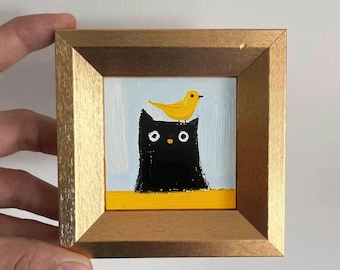 Puede incluir: Pintura enmarcada que presenta un gato negro con un pájaro amarillo posado en la cabeza. La obra de arte tiene un fondo azul claro y una línea horizontal amarilla en la parte inferior. El marco es de madera con un borde interior dorado.