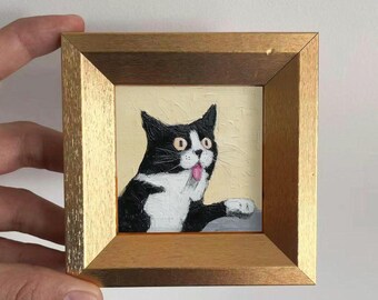 May include: A framed painting of a black and white cat with its tongue sticking out. The cat is in a gold-colored wooden frame. The cat's eyes are wide open, and it appears surprised. The background is a light yellow color.