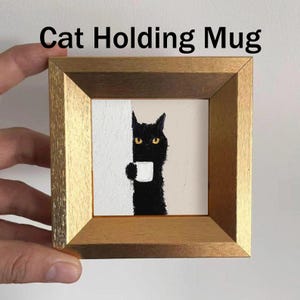 May include: A framed artwork featuring a black cat holding a white mug. The cat has yellow eyes and is set against a neutral background. The frame is a gold color, and the text "Cat Holding Mug" is at the top.
